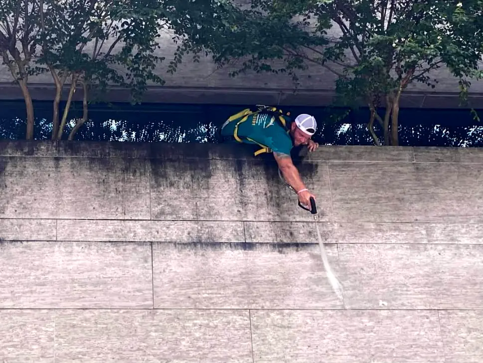 Harnessed crew member leaning over a parapet to pressure wash black streaks off a stone facade