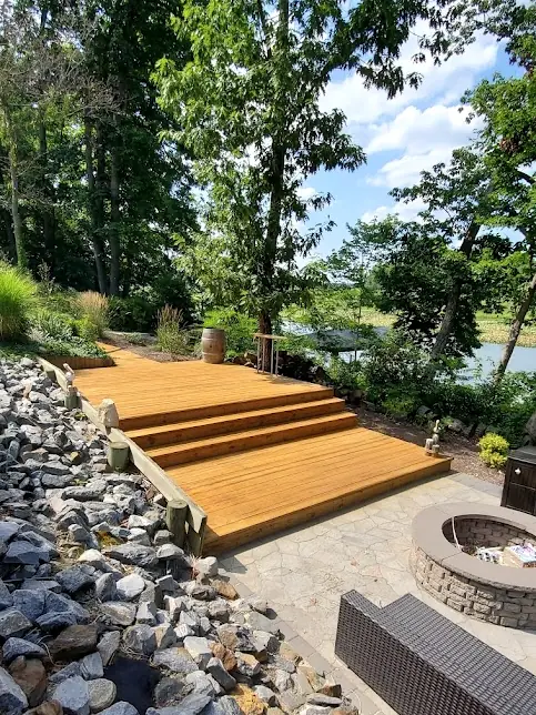 Wide stained deck steps beside a stone patio and fire pit near the water