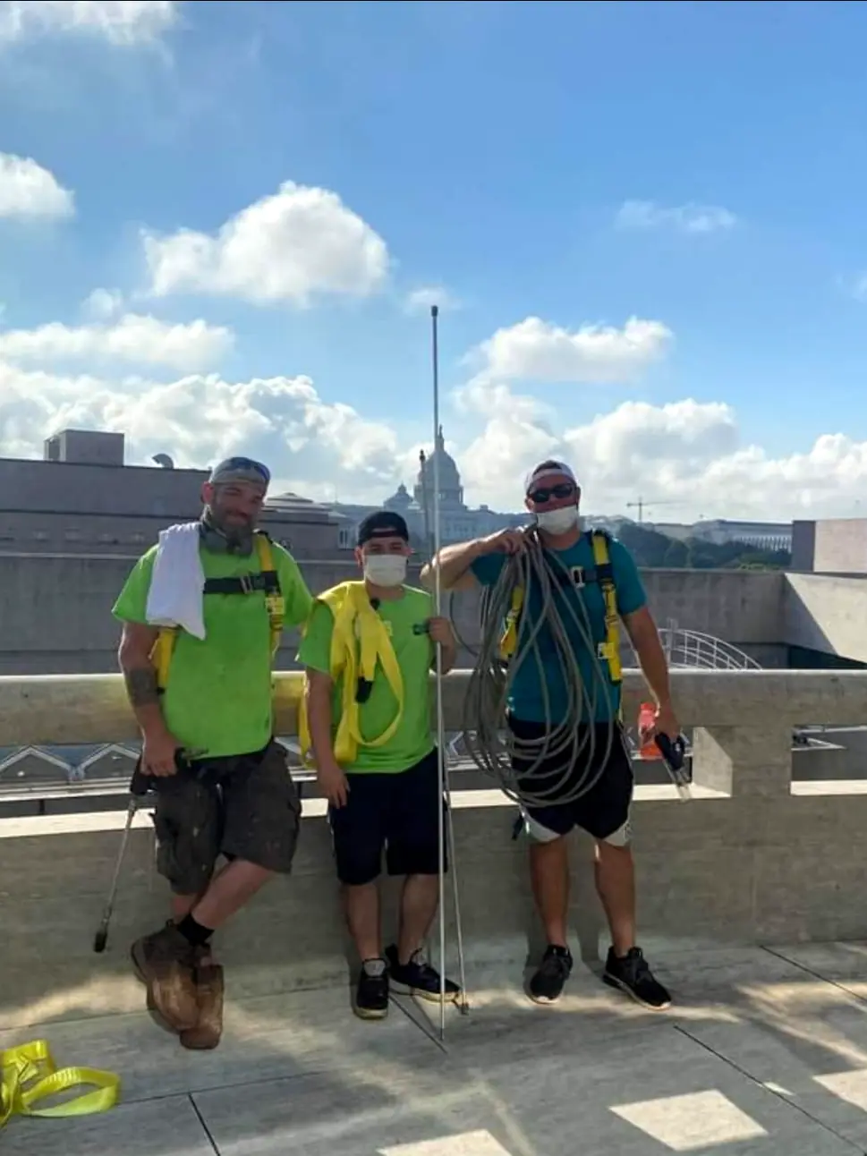 Three Ultimate Powerwash crew members in fall-protection harnesses on a commercial rooftop, with hose and wand
